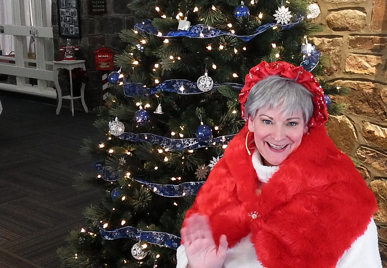 An image of Santa Sue smiling and
        waving while sitting in front of a Christmas tree